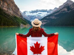 woman wearing red and black checkered dress shirt and beige fedora hat holding canada flag looking at lake