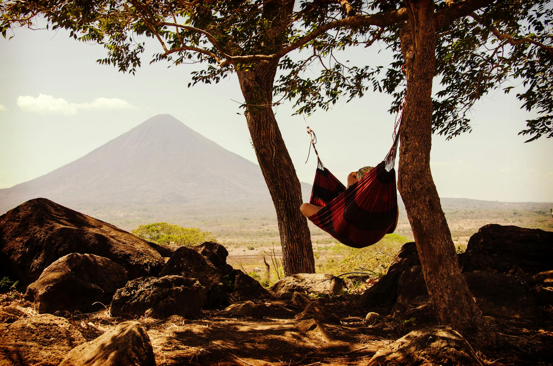person lying on black and red hammock beside mountain under white cloudy sky during daytime