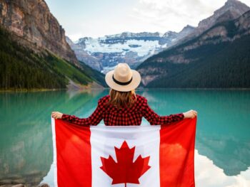 woman wearing red and black checkered dress shirt and beige fedora hat holding canada flag looking at lake