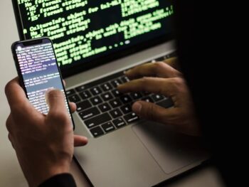 unrecognizable hacker with smartphone typing on laptop at desk